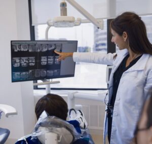Dentist pointing at dental X-rays on a screen while discussing tooth decay and cavity detection with a patient in a modern dental office.